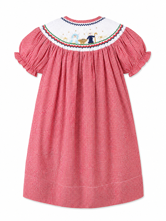 A girl's bishop red and white gingham dress shown from the back, featuring a white smocked yoke with colorful smocked nativity scene and a red, blue, and green border.